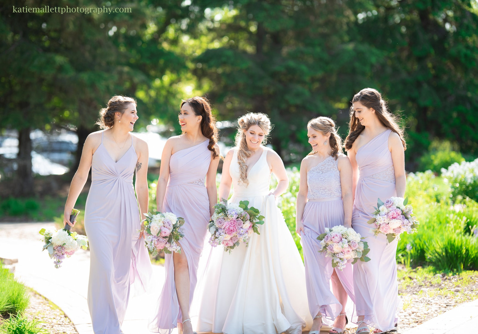 A bride and her bridesmaids in lavender dresses walking together on a garden path.