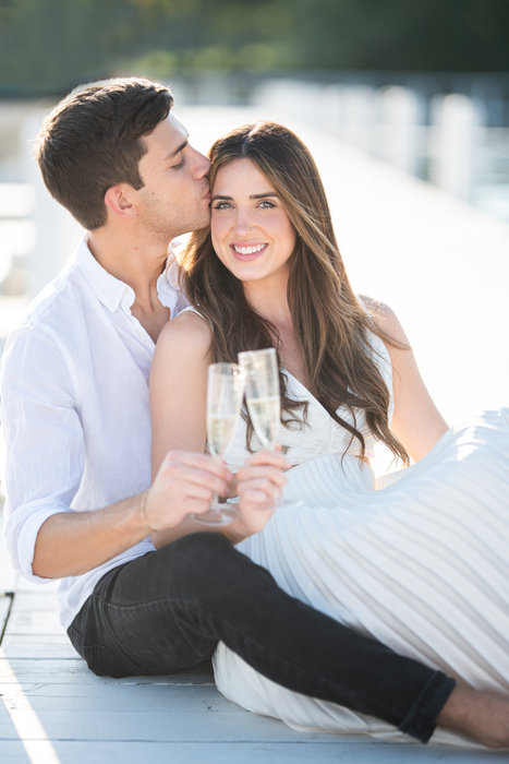 An engaged couple sitting close together, laughing as he kisses her cheek.