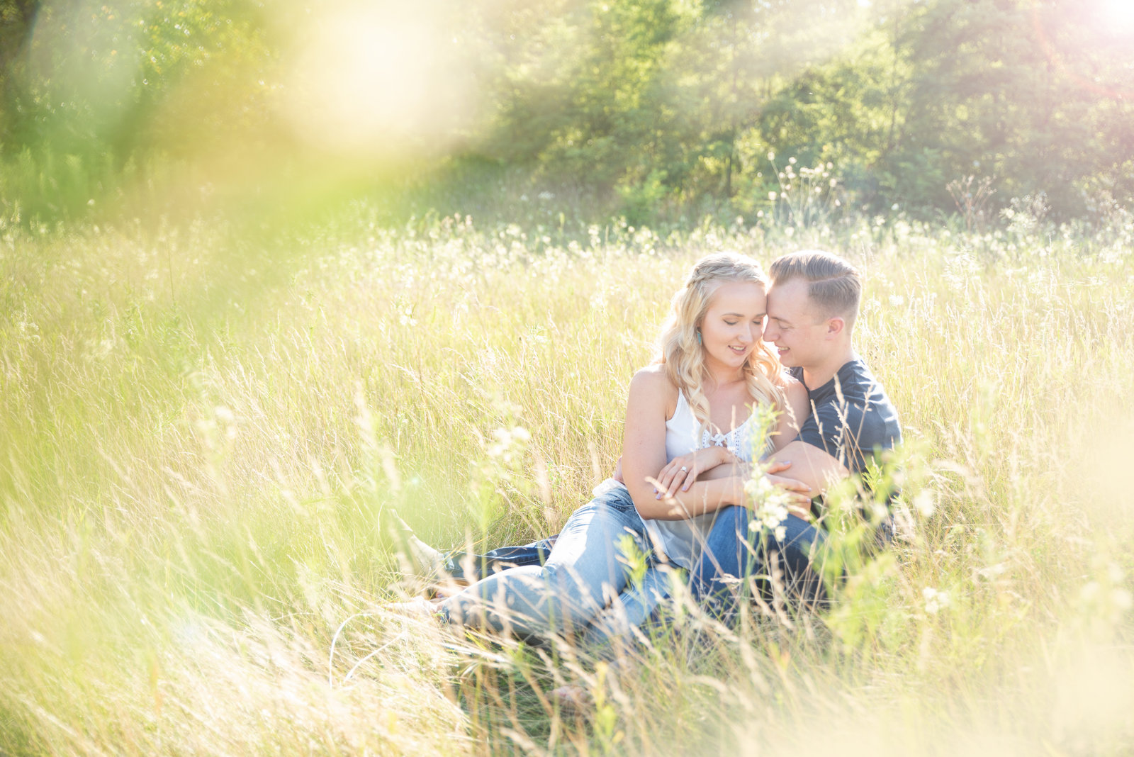 A couple sitting close together in tall meadow grass, with sunlight behind their heads.