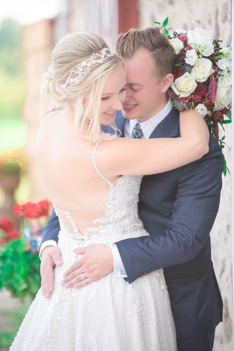 Karley and Chris smiling close together, with a pale pink bouquet and sunlight in the foreground.