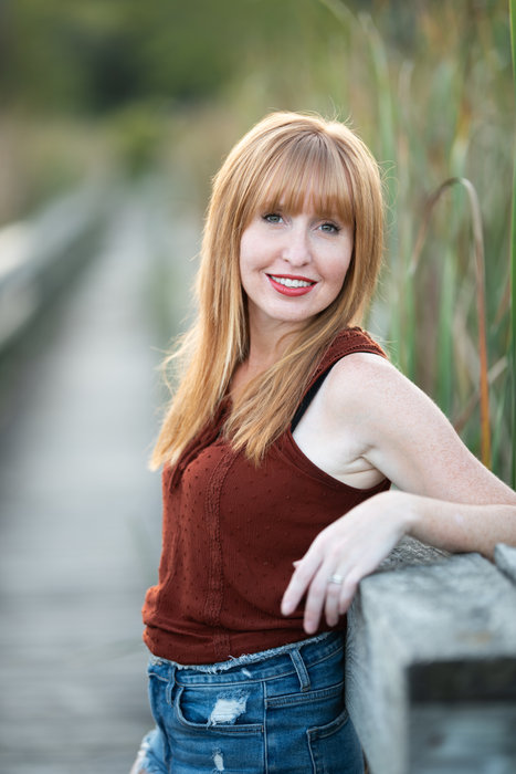Katie Mallett, with red hair and a rust-colored top, leaning on a wooden boardwalk railing.
